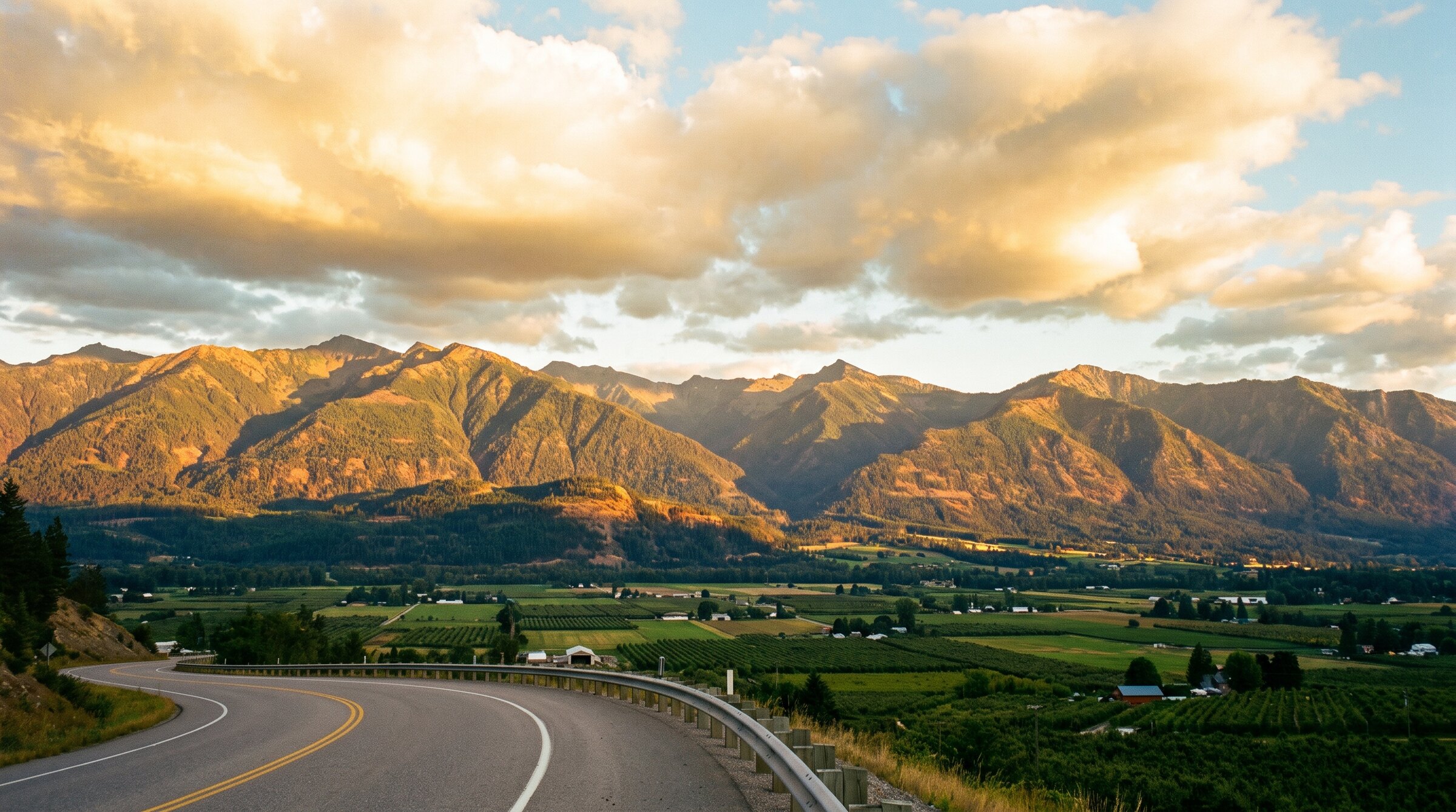 Highway 3 sweeping down into the Creston valley — orchards and farmland below gold-lit Kootenay peaks at sunset